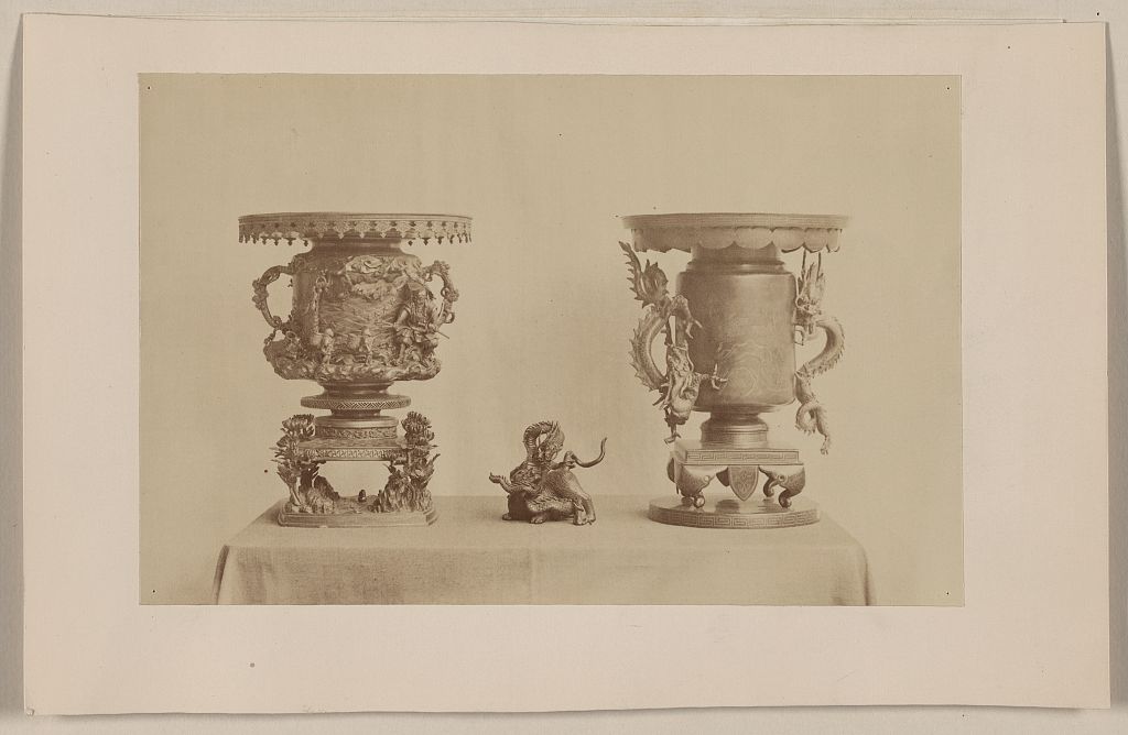 A picture of Tall, metal, ornate plant stands and figurine, all with dragon motif, on a table